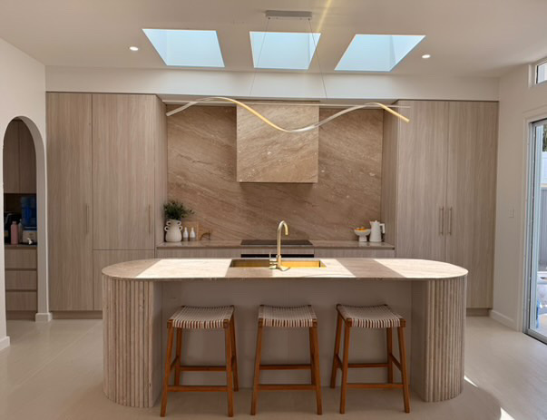 Kitchen with a curved fluted island, stone splashback and rangehood under three skylights